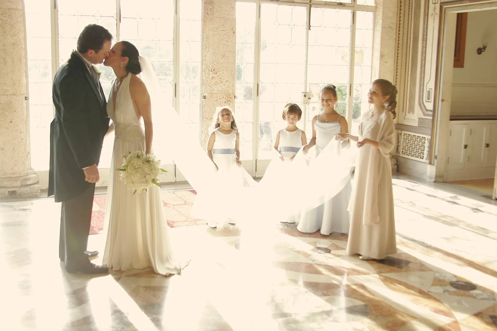 Newlyweds share a kiss in a sunlit Vizcaya salon as flower girls look on